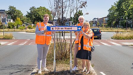 Naast een blauw bewegwijzeringsbord staan drie personen in oranje veiligheidshesjes.