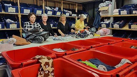 Vier vrijwilligers zijn de binnengekomen kleertjes aan het selecteren achter grote rode bakken.