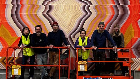 Een groep mensen staat voor een kleurrijke muurschildering. Onder hen de twee kunstenaars die in het artikel worden genoemd.