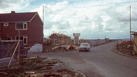  Een historische foto van de wijk Barnewiel in Oudkarspel tijdens de bouw in de jaren ‘70. Op de voorgrond is een deels afgebouwde woning te zien, omringd door bouwmaterialen. Een klassieke auto rijdt over een onverharde weg, terwijl op de achtergrond meerdere huizen in aanbouw zijn met steigers en houten constructies.