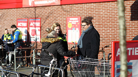 Burgemeester staat voor een supermarkt. Hij is in gesprek met een politieagent en een bewoner.