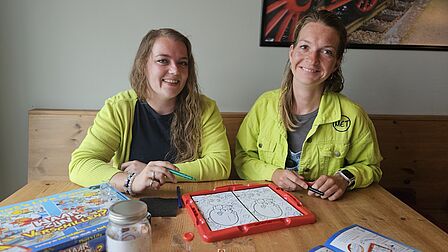 Op de foto zijn Celine en Jacky te zien die zitten aan de eettafel met spelletjes op tafel.
