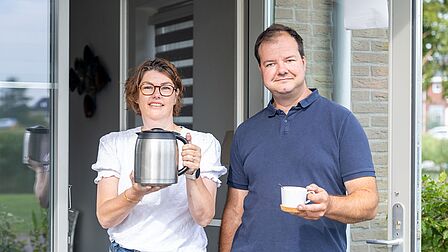 Op de afbeelding staan Daniel en Willemein bij hun voordeur met een koffiekan in handen en lachen ze richting de camera. 