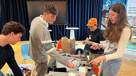 Jonge studenten staan rond een tafel en werken samen aan opdrachten.