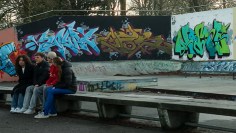 Op deze header zie je een lange betonnen bank met daarop drie jongeren. Achter hen zijn borden met grafitti zichtbaar met diverse teksten in licht- en donkerblauw en groen. Lastig leesbaar ...