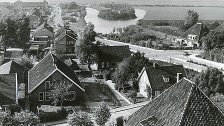 Een zwart-wit luchtfoto van de wijk Barnewiel in Oudkarspel na de verkaveling en nieuwbouw. Op de voorgrond staan traditionele huizen met puntdaken en tuinen. Een rechte weg loopt door de wijk, met huizen aan beide zijden. In de achtergrond is een waterplas met een klein eiland te zien, omringd door groen en open landschap. De foto toont de overgang van een landelijke omgeving naar een meer bebouwde woonwijk.