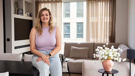 Er zit een dame (Pauline) op een eettafel in de woonkamer. Ze lacht richting de camera.
