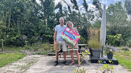 Inwoners Jerry en Cindy, woonachtig in De Noord, staan in hun groene tuin met de gewonnen gouden gieter in hun hand en een tuinbon ter waarde van 100 euro. Op de achtergrond is de groene tuin te zien, net als de blauw lucht en een ingeklapte parasol.