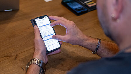 Een man zoomt met zijn vingers in op het scherm van zijn smartphone om een document beter te lezen. Naast hem op tafel ligt een pasjeshouder en de warmtecamera van energiecoach Peter